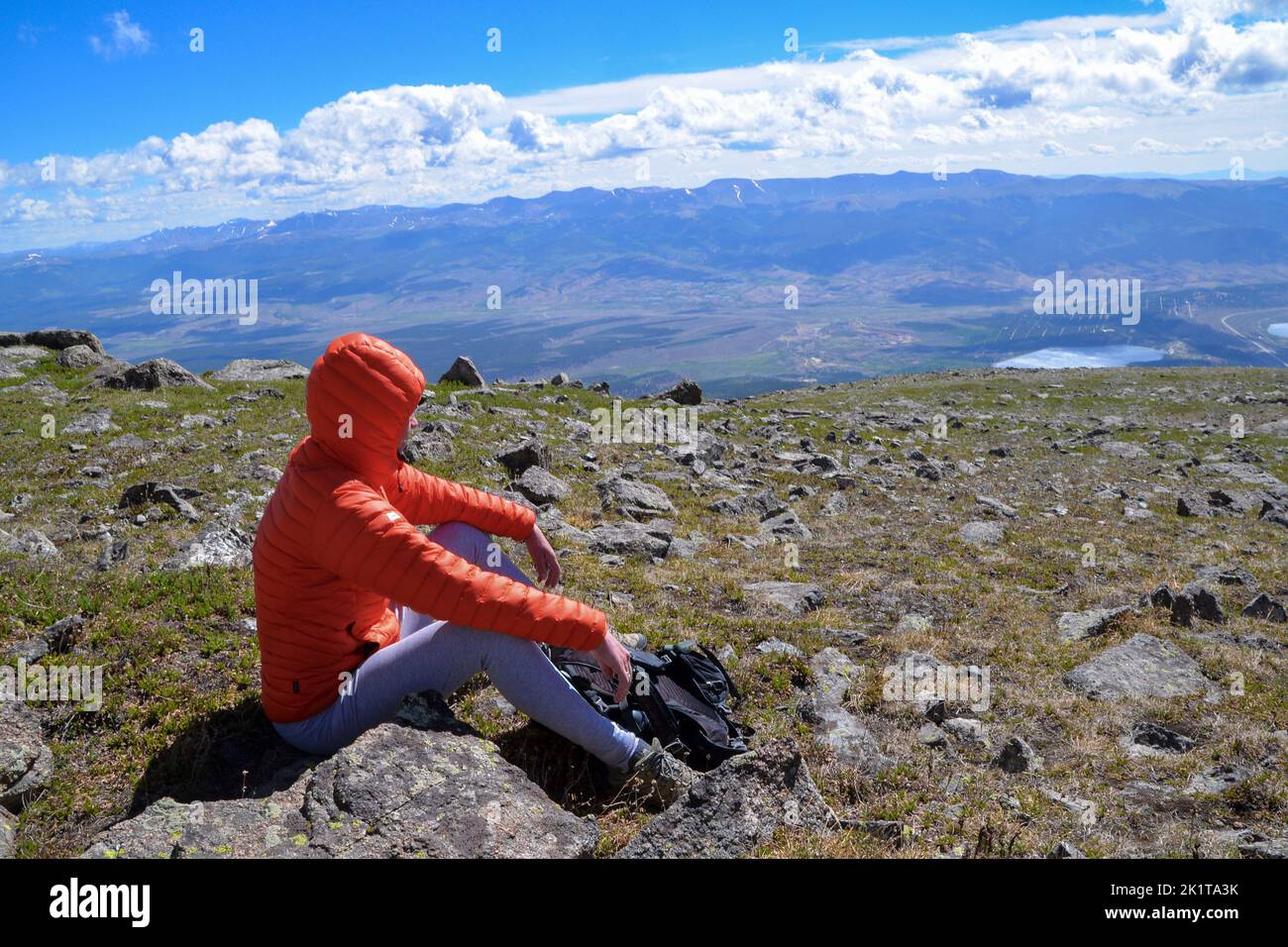 Eine Frau in einer orangefarbenen Jacke mit Kapuze sitzt für eine Pause mit Blick auf die Berge nahe der Spitze des Mount Elbert, dem höchsten Gipfel in Colorado USA Stockfoto
