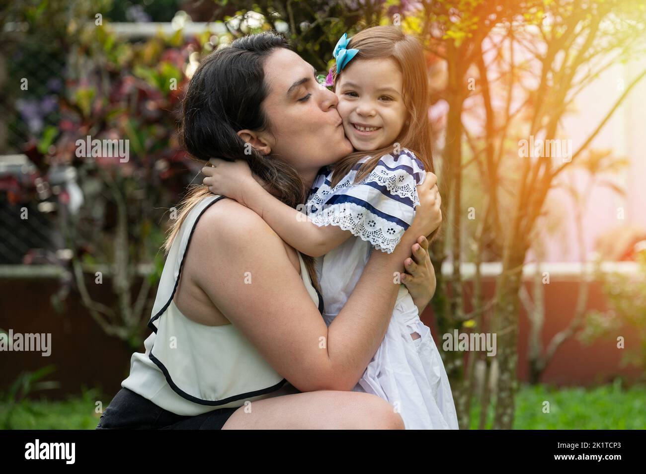 Hispanische Mama küsst ihre Tochter auf sonnigem Parkhintergrund Stockfoto