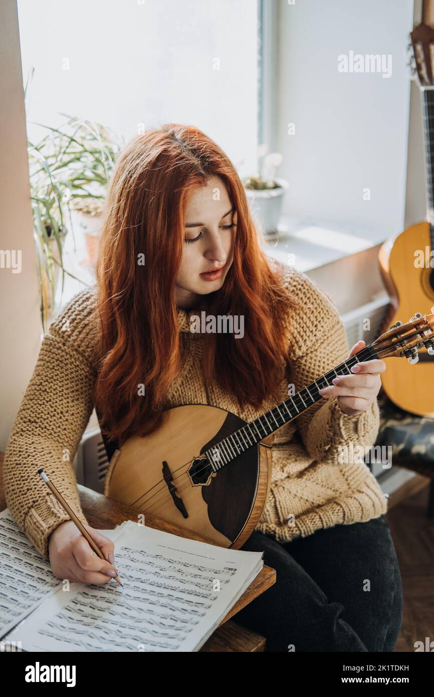 Redhead Frau spielt akustisches Domra Folk Musical Instrument und schaut in die Note. Junge Frau, die zu Hause Musikunterricht nimmt Stockfoto