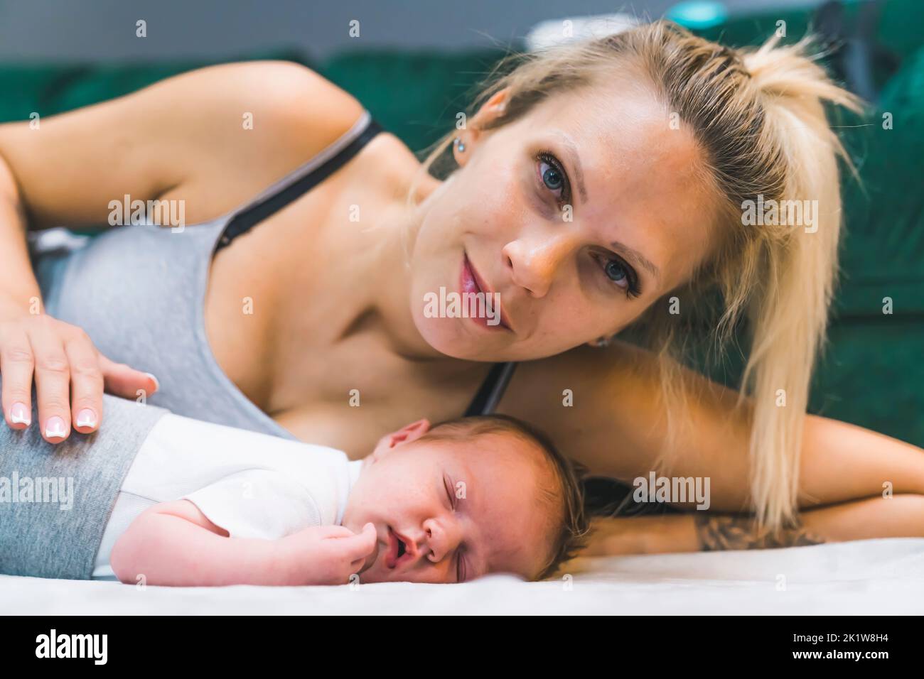 Weiße blonde Frau, die auf ihrer Seite auf dem Boden liegt und sich mit ihrem neugeborenen Jungen kuschelt und mit einem Lächeln in die Kamera schaut. Stolze, liebevolle Mutter. Horizontale Innenaufnahme. Hochwertige Fotos Stockfoto