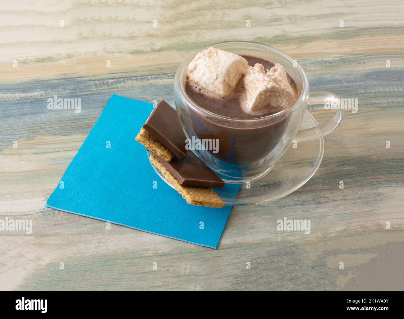 Heiße Schokolade in durchsichtiger Tasse mit schmelzenden Marshallows und Schokolade auf graham Crackers Snack auf Untertasse auf blauer Serviette Stockfoto