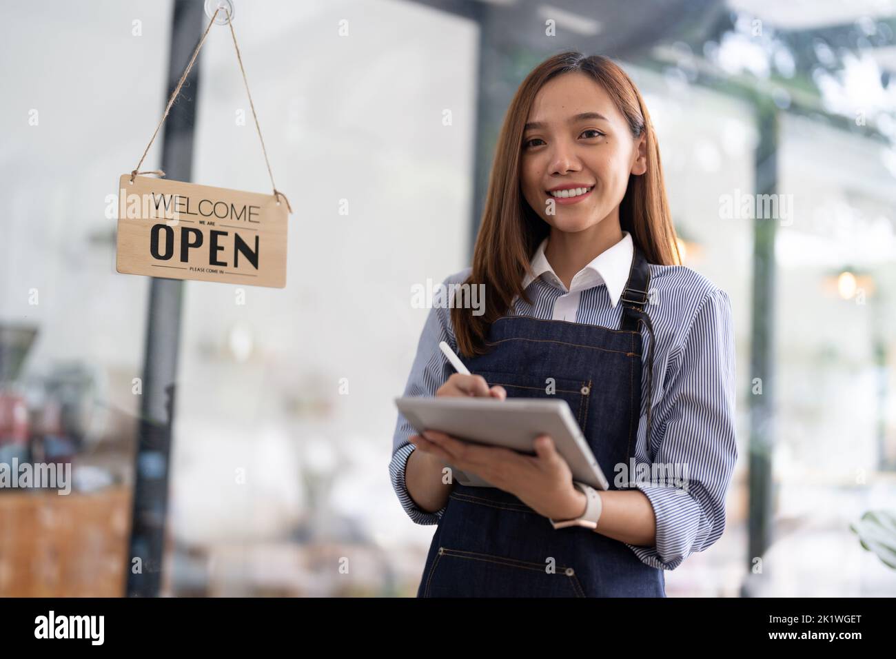 Charmante asiatische Geschäftsfrau in einer Schürze hält ein Auftragsbuch vor einem Café-Schalter mit einem strahlenden Lächeln bereit, um zu öffnen und Aufträge entgegennehmen. Stockfoto