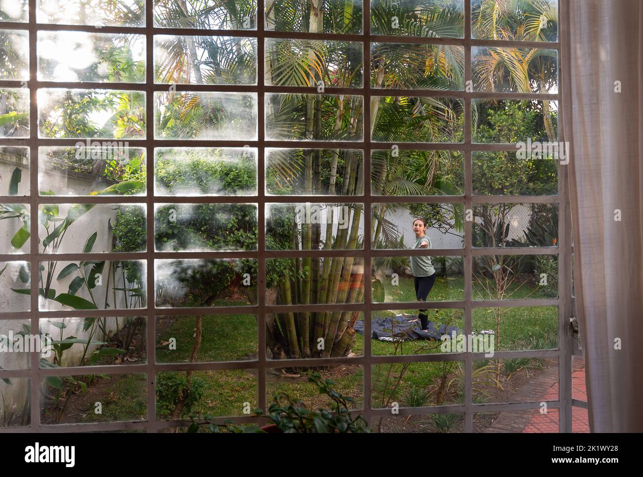 Frau praktiziert Yoga im Hinterhof, umgeben von Pflanzen und Bäumen, eingefangen durch das Fenster des Hauses mit tropischen Vibes und Palmen Stockfoto