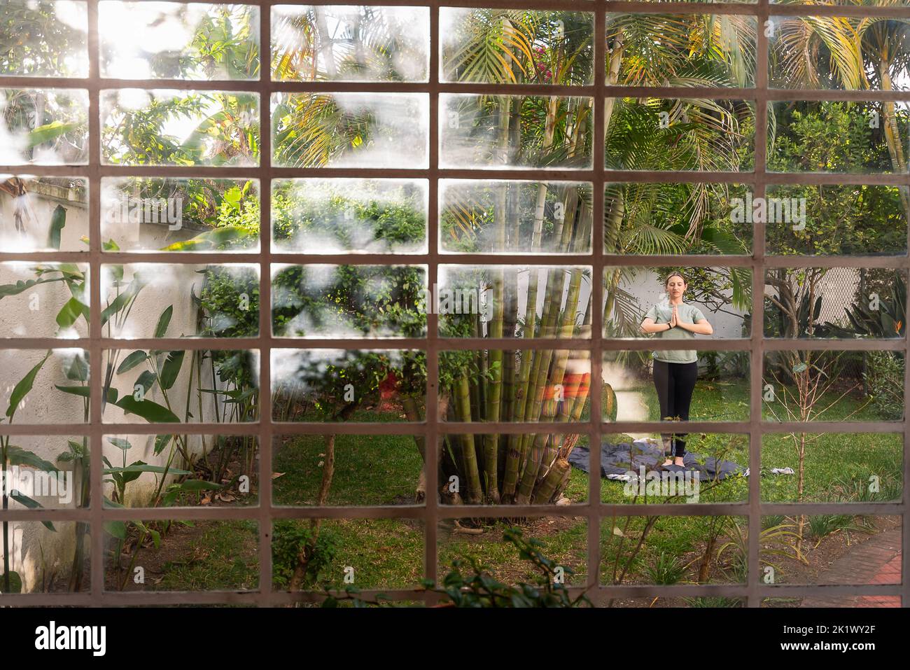 Frau praktiziert Yoga im Hinterhof, umgeben von Pflanzen und Bäumen, eingefangen durch das Fenster des Hauses mit tropischen Vibes und Palmen Stockfoto