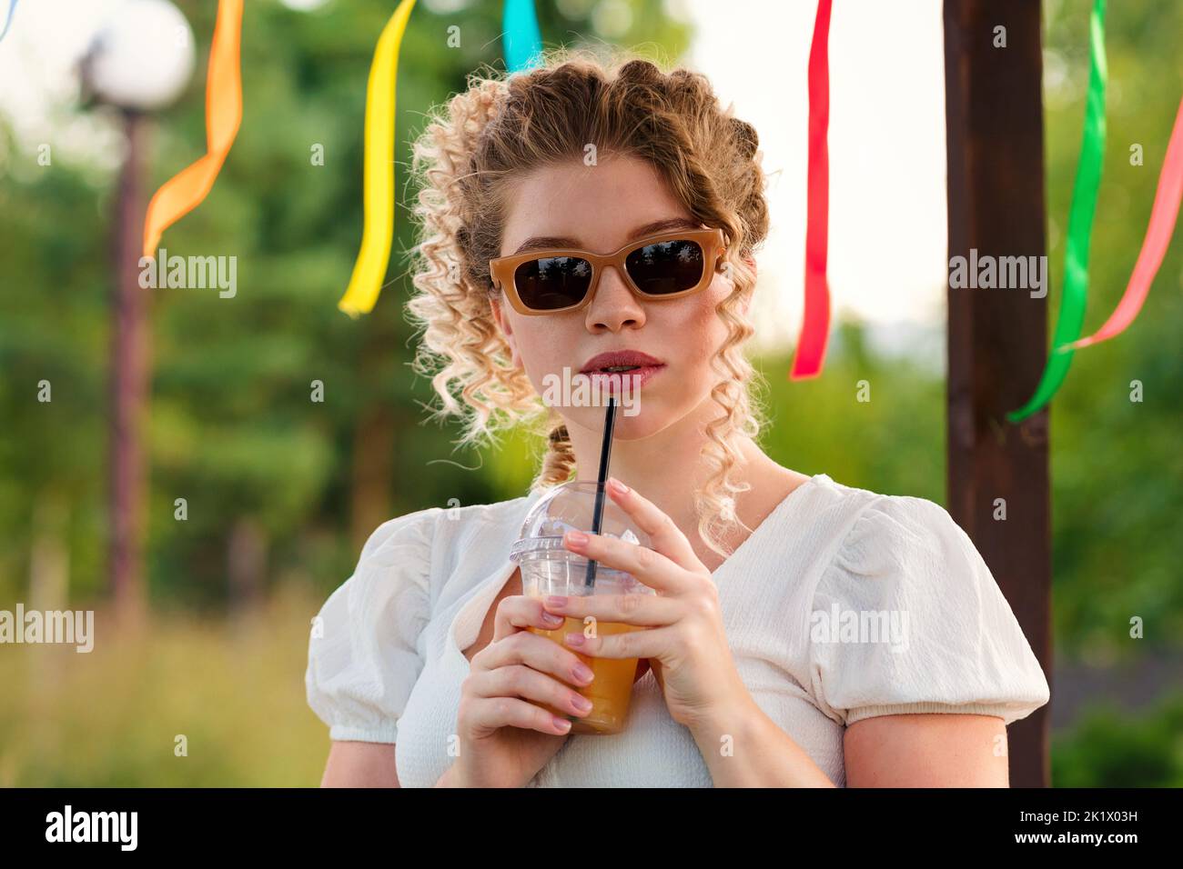 Attraktive blonde lockige Mädchen in Sonnenbrillen und Sommer-Outfit genießen Limonade, alkoholfreien Cocktail auf Party im Stadtpark auf offener Veranda. Du bist süß Stockfoto