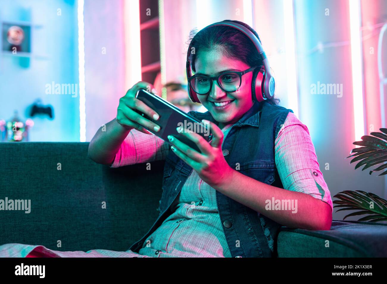Aufgeregt Mädchen spielen Videospiel auf Handy mit schnurlosem Headset auf Sofa zu Hause auf Neon-Hintergrund - Konzept des Cyberspace, Hobby und Stockfoto