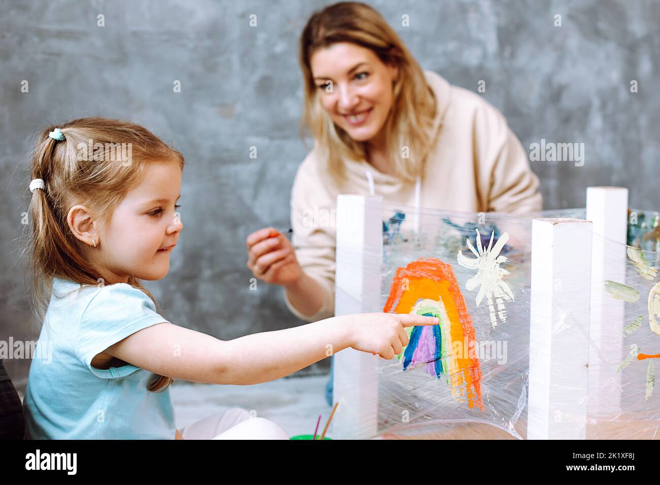 Lehrerin mit blondem Mädchen, das Regenbogen auf Öltuch malt. Spaß beim Lernen in Kinderentwicklungszentrum Stockfoto