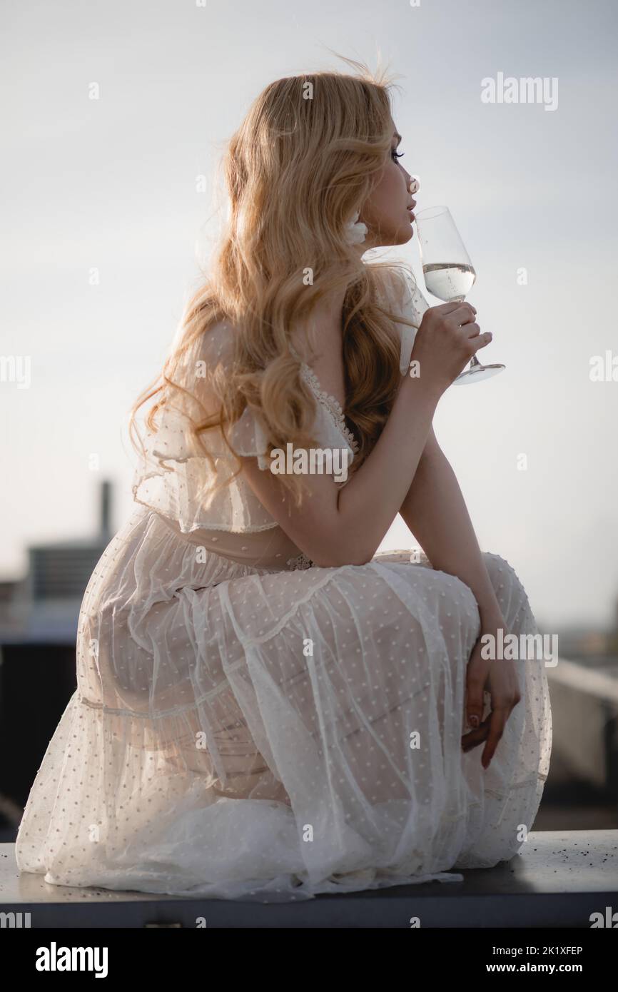 Schöne Frau in romantischem weißen Kleid hockt sich seitlich mit einem Glas Champagner auf der Dachterrasse und schaut weg. Elegante Dame im stilvollen Outfit und Stockfoto