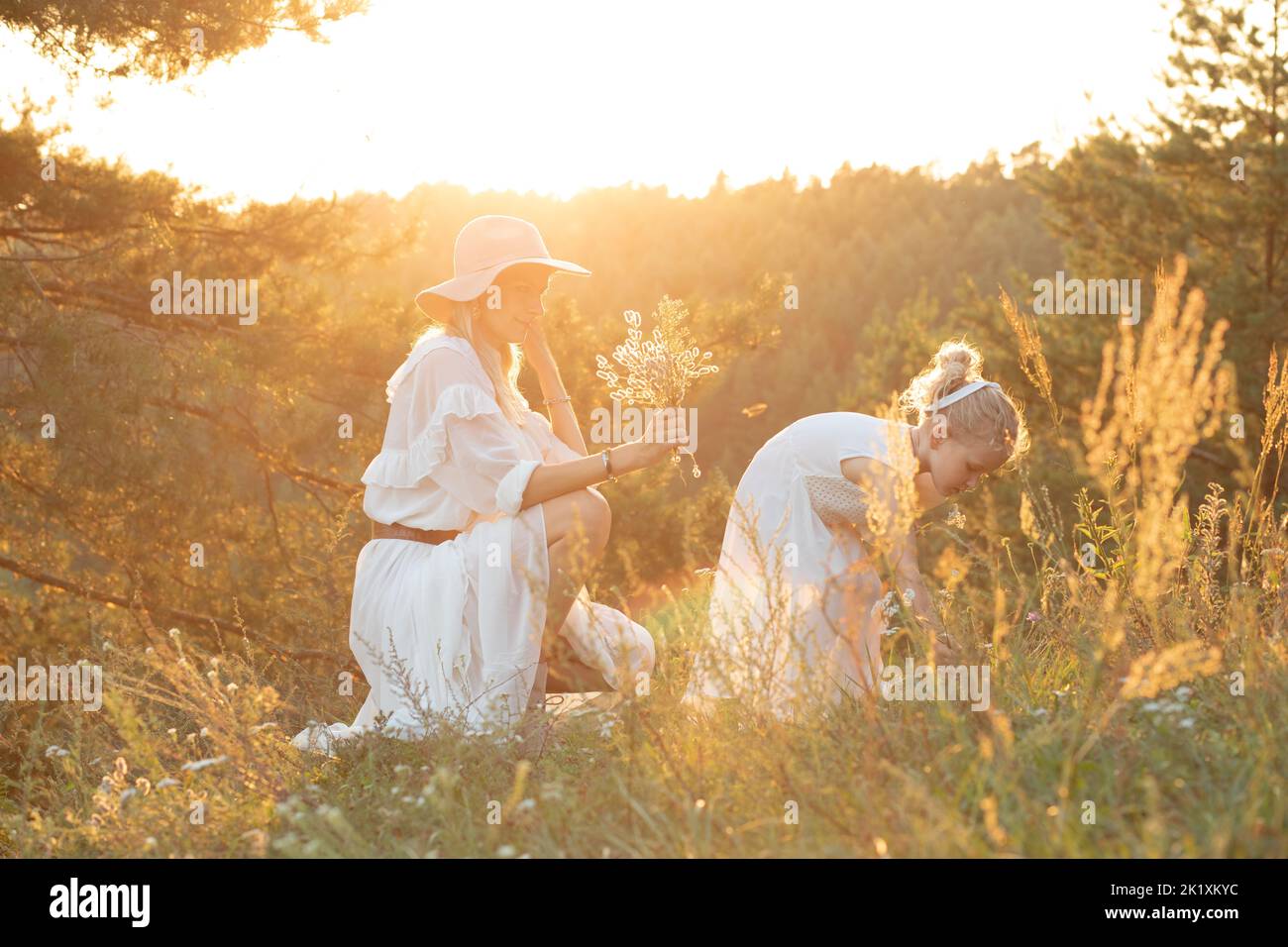 Lächelnde, fröhliche, fröhliche, ruhige Frau, Mutter im Hut und kleines Mädchen in weißem Kleid haben Spaß und sammeln Kräuter in der Natur Stockfoto