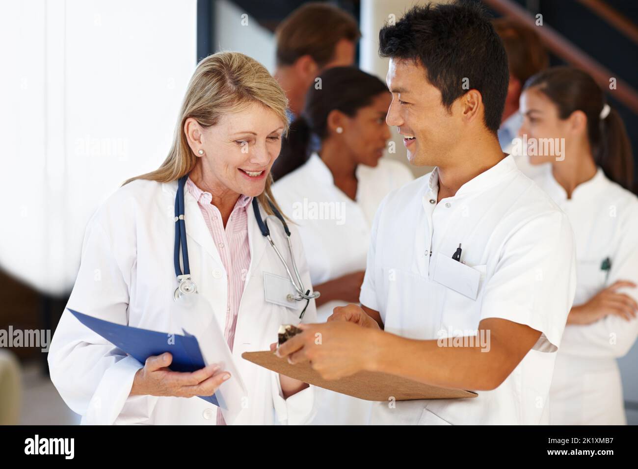 Sie lernen ziemlich schnell. Zwei Mediziner schauen sich ein Clipboard mit einer Reihe von Kollegen im Hintergrund an. Stockfoto