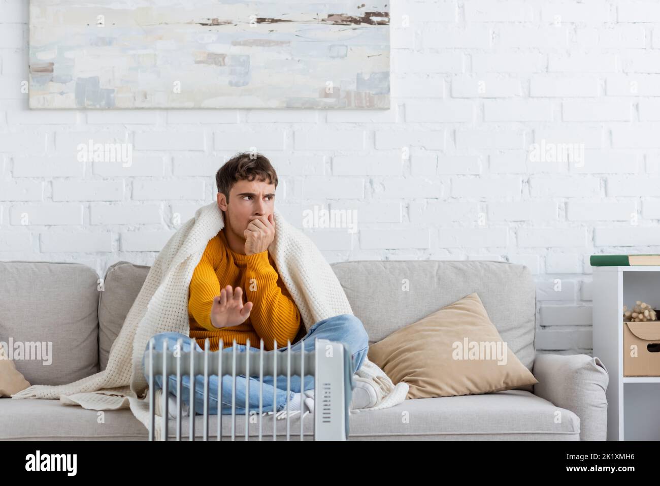 Junger Mann, der mit einer Decke bedeckt ist, sitzt auf der Couch und wärmt die Hände in der Nähe des modernen Heizkörperheizers, Stockbild Stockfoto
