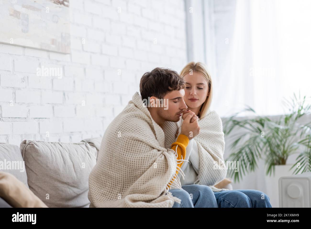 Junger Mann wärmende Hand der Freundin in Decke im Wohnzimmer sitzend bedeckt, Stockbild Stockfoto