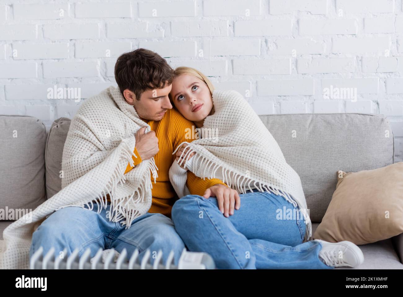 Junges Paar mit Decke bedeckt, das auf der Couch sitzt und sich in der Nähe des modernen Heizkörperheizkörpers wärmt, Stockbild Stockfoto