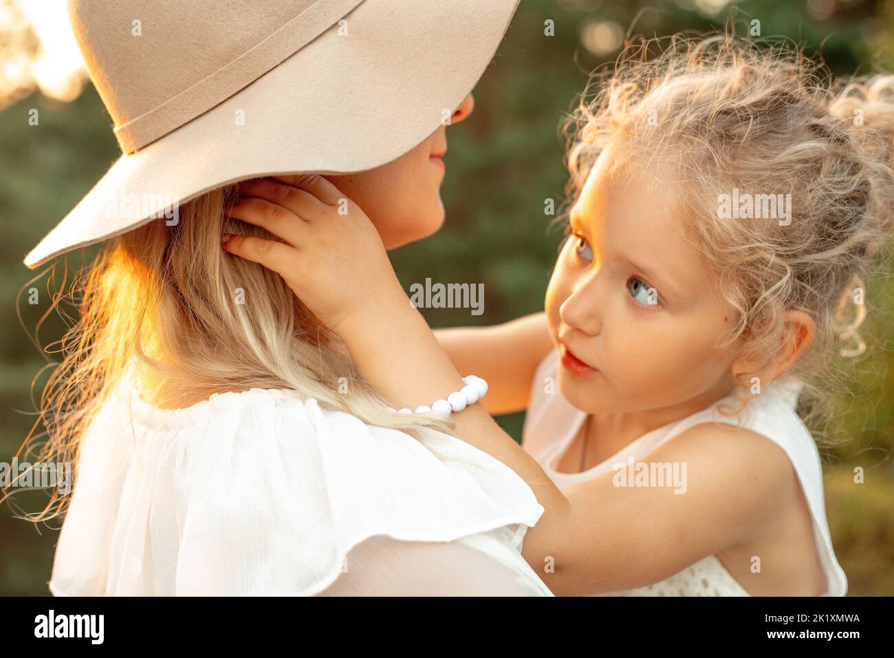 Unwiedererkennbar helle, lächelnde Frau in Hut umarmt kleines blondes Mädchen, Tochter mit Armband auf natürlichem unscharfen Hintergrund Stockfoto