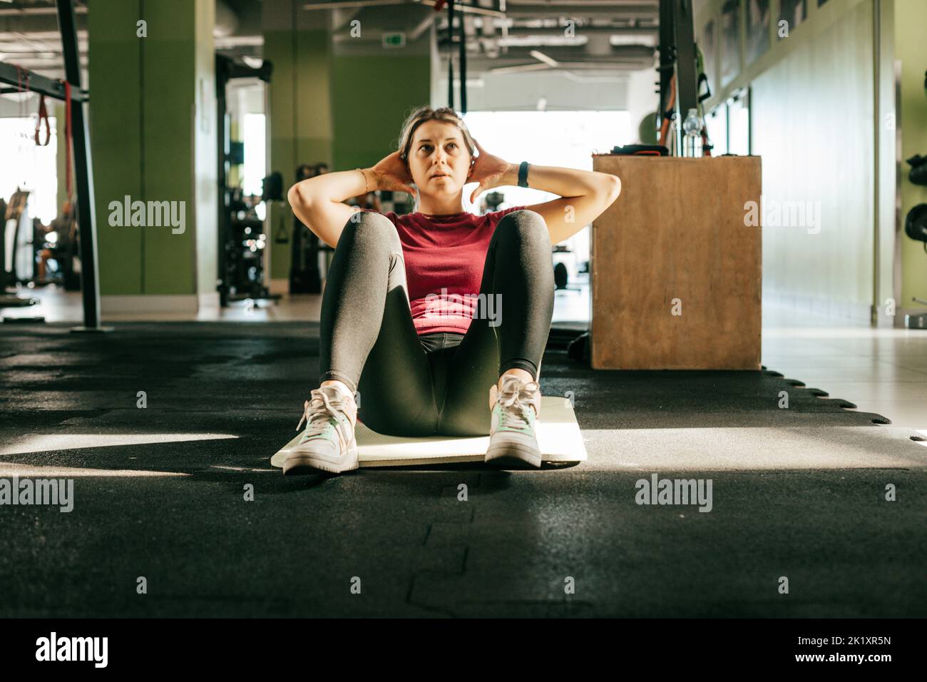 Übergewichtige Mädchenpumpenpresse im Fitnessstudio, das auf einer Matte sitzt, mit Händen auf dem Kopf und gebeugten Knien. Junge Frau tun Cardio-Training, indem sie Körper. Gewichtsverlust Stockfoto