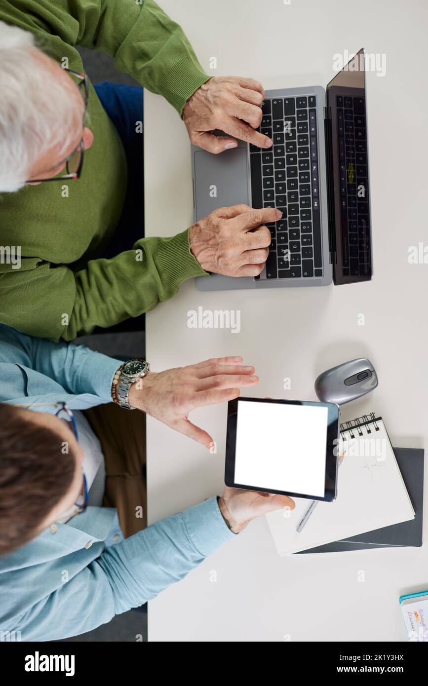 Der junge Pädagoge zeigt dem Studenten, wie er ein Problem auf seinem Tablet beim Projekt lösen kann, während der Schüler auf einem Laptop tippt. Stockfoto
