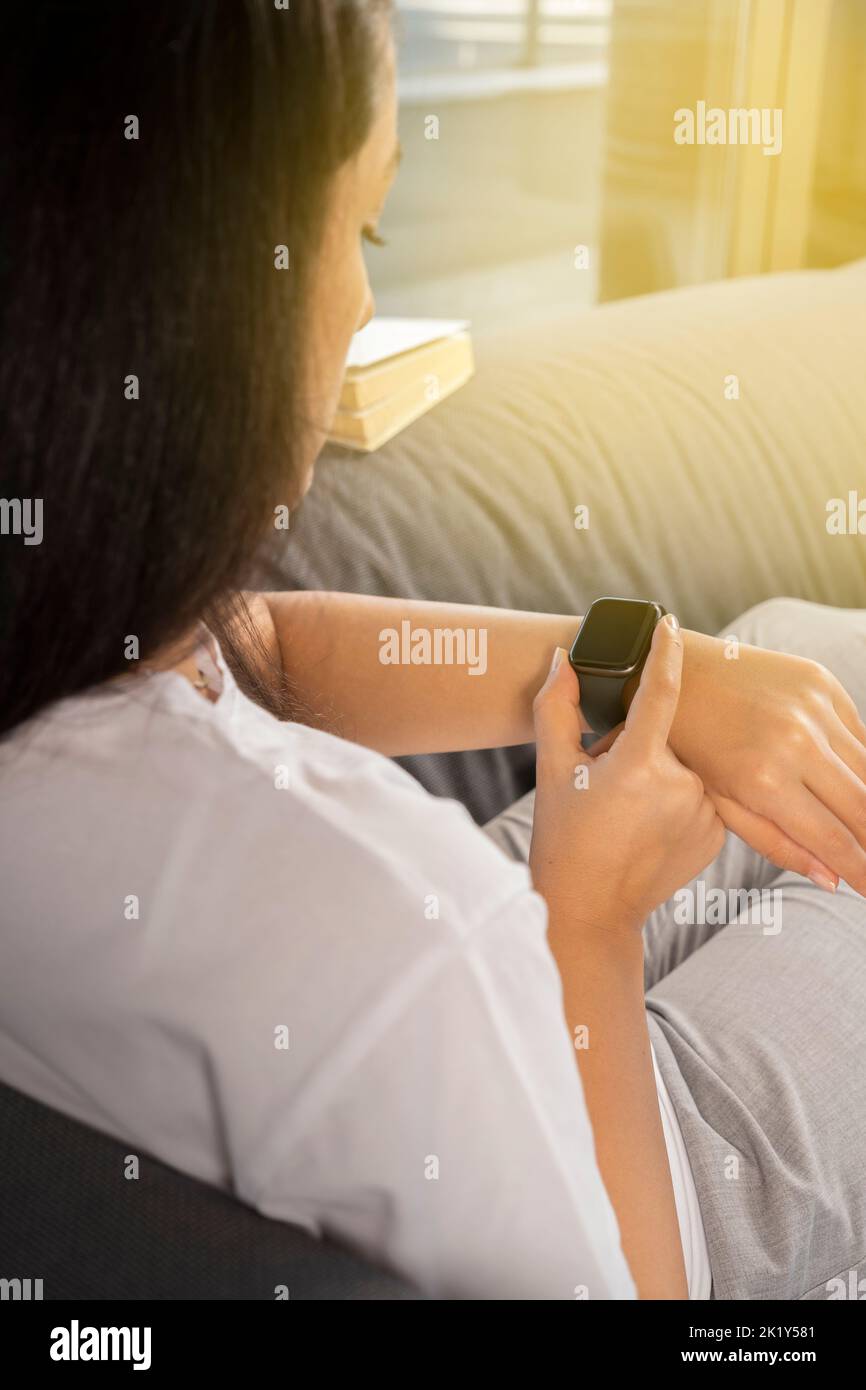 Mit Smartwatch, über die Schulter der jungen Frau mit Smartwatch. Zu Hause sitzen, auf der Couch entspannen. E-Mails, Nachrichten und Herzschläge werden überprüft. Stockfoto