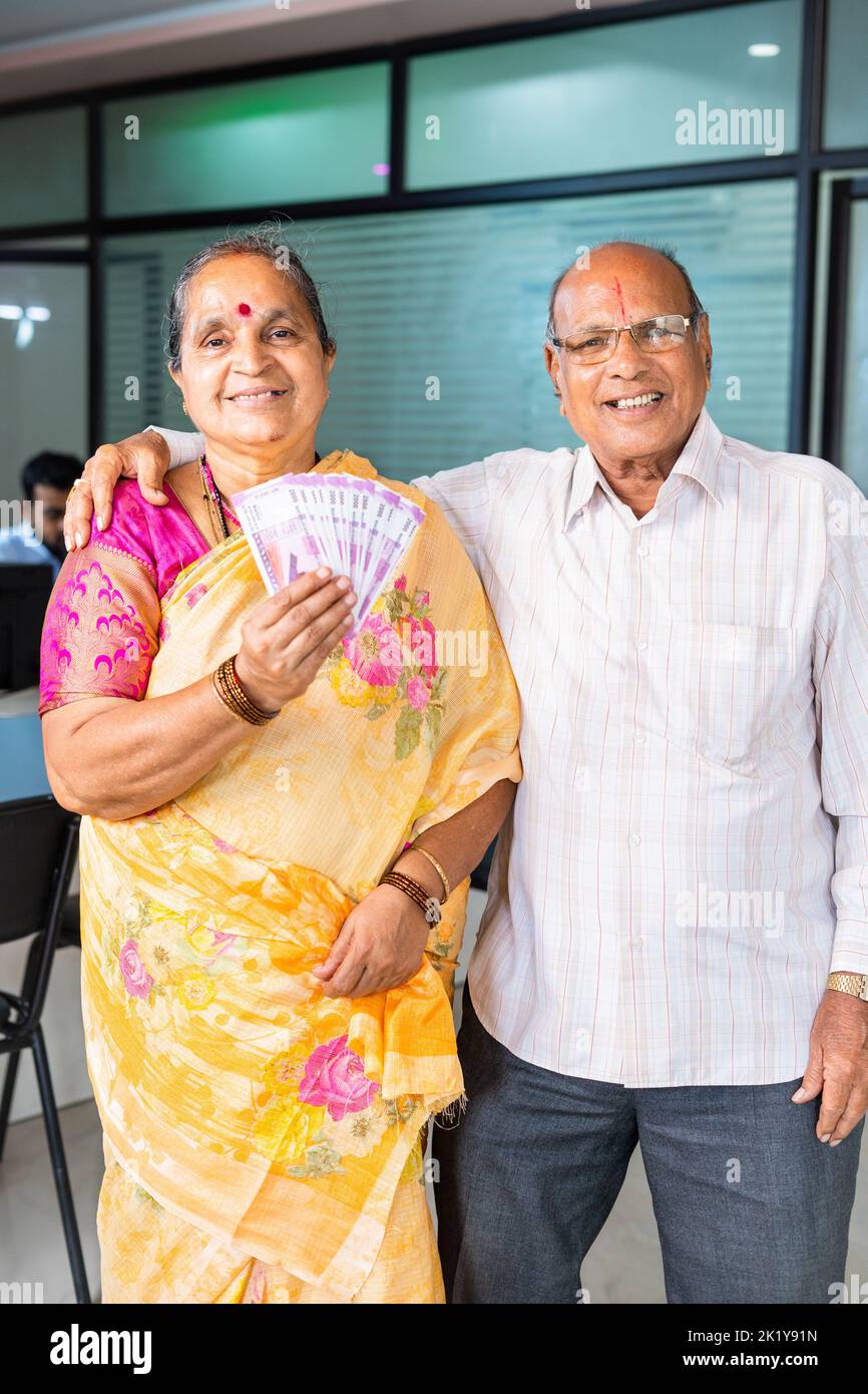 Vertikale Aufnahme eines glücklichen lächelnden älteren Paares, das indisches Geld oder Geldscheine zeigt, indem man die Kamera auf die Bank schaut - Konzept der Kreditgenehmigung, Investition Stockfoto