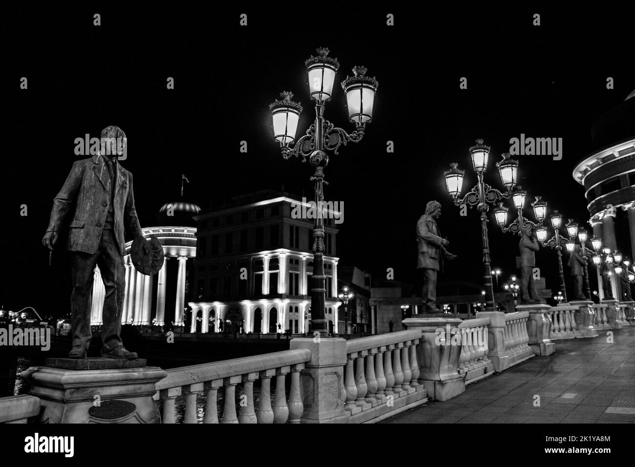 Statuen, die die Brücke der Kunst schmücken, über den Vardar-Fluss, Skopje, Nordmakedonien. Nachts mit Lampenpfosten, die die Brücke erleuchten. Stockfoto