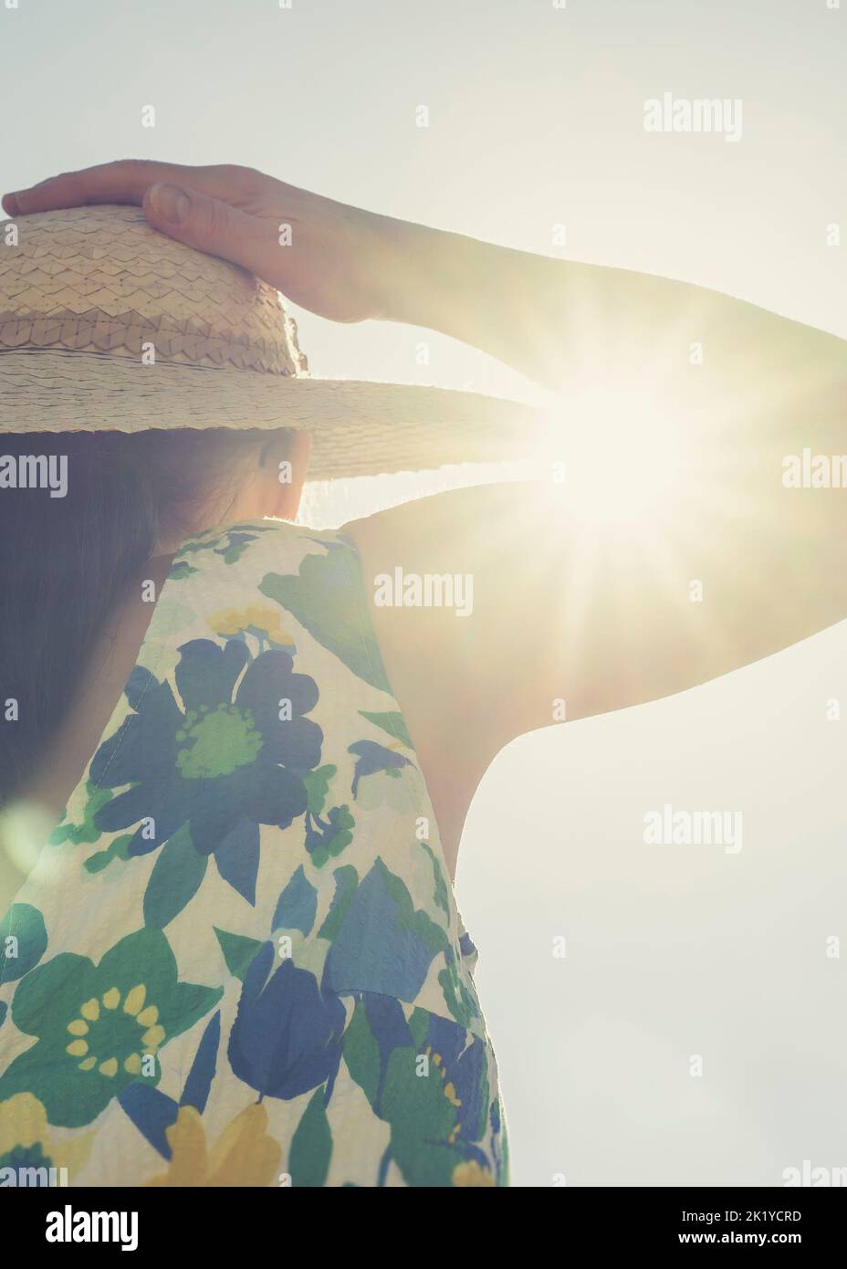 Nahaufnahme einer Frau in einem Sommerkleid, die sich an ihrem Strohhut auf ihrem Kopf festhält, wobei der intensive Sonneneinbruch durch die gekrümmte Armfalte bricht. Stockfoto