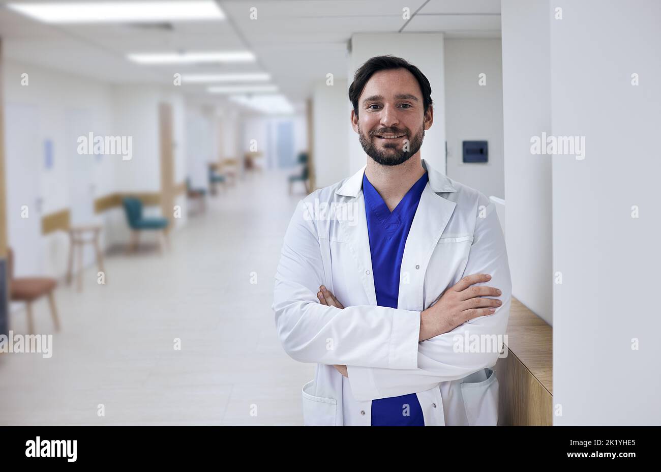 Porträt eines glücklichen männlichen Arztes, der mit gekreuzten Armen im Krankenhausflur steht. Gesundheitsfachkraft Stockfoto