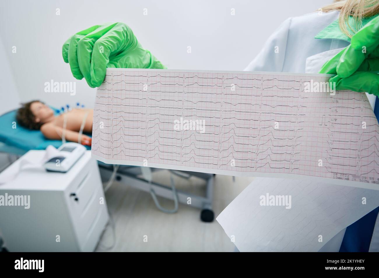 Kardiogramm-Test des kindlichen Herzens in den Händen des Arztes vor dem Hintergrund eines jungen Patienten, der auf dem medizinischen Sofa mit Vakuumsonden im Herzbereich liegt Stockfoto