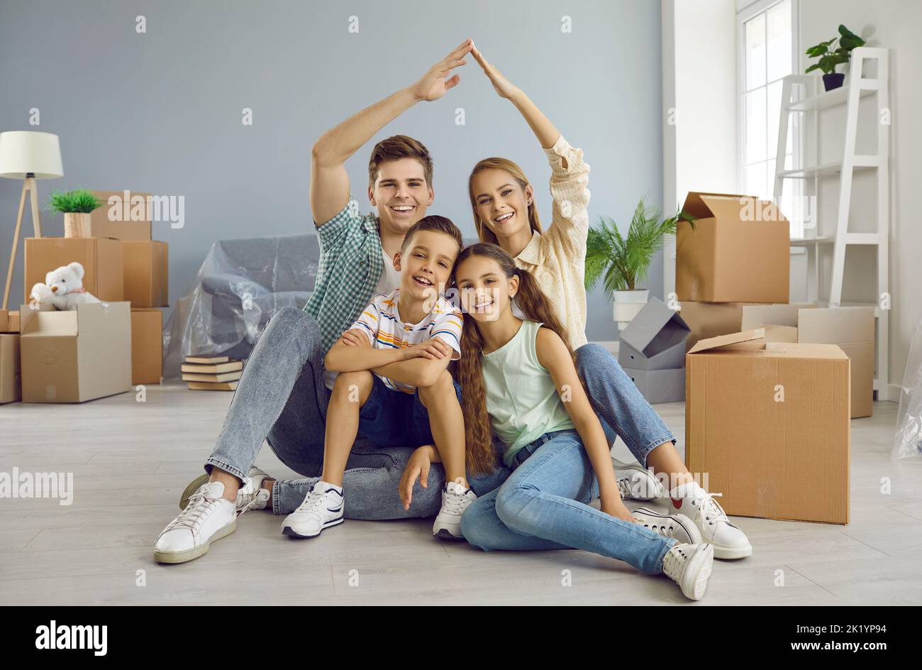Glückliche Familie mit Kindern, die am Umzugstag unter einem symbolischen Dach in ihrem neuen Haus sitzen Stockfoto