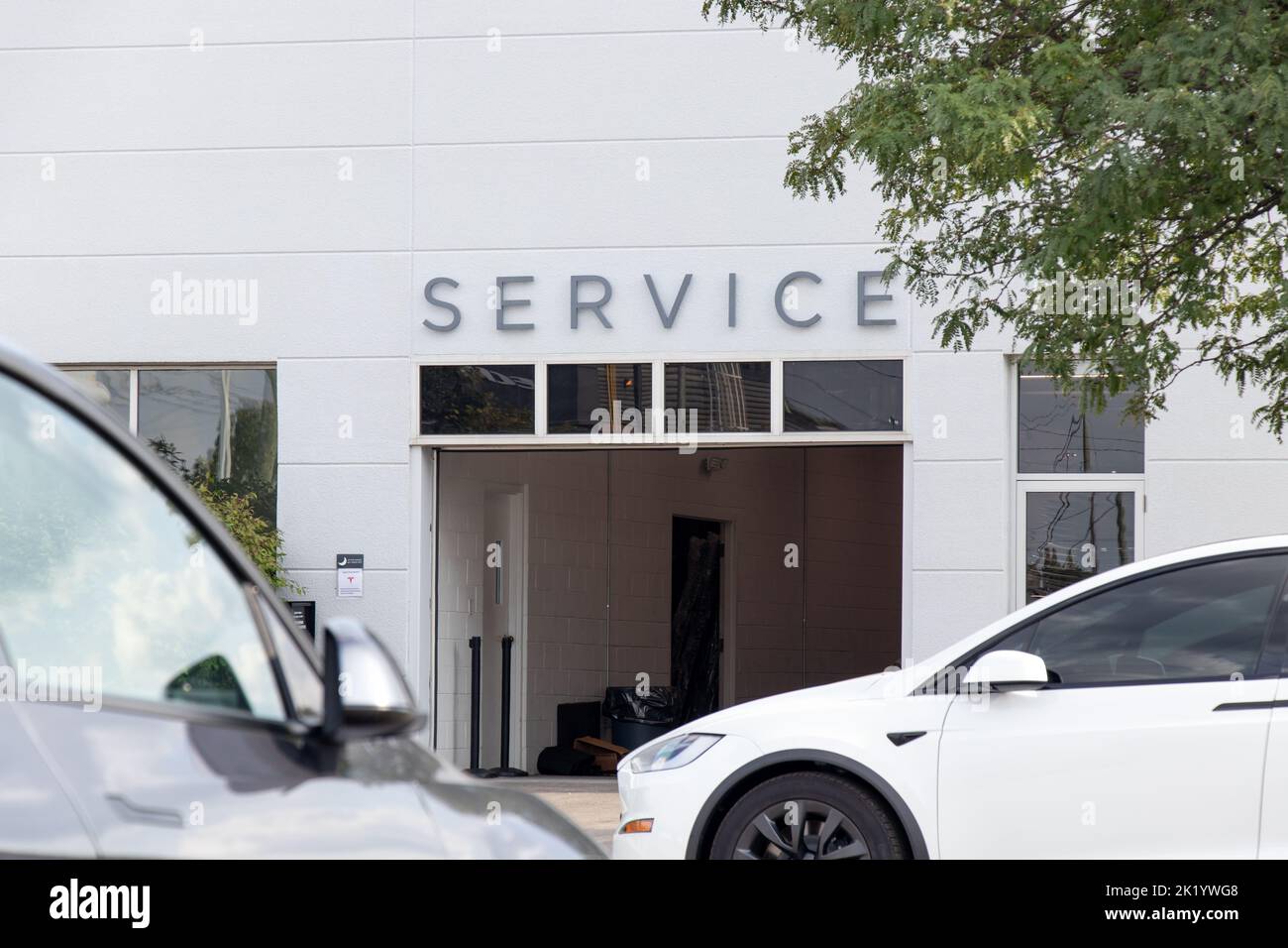 Über dem Eingang einer Garage zu einem Tesla-Händler ist ein Service-Schild zu sehen; geparkte Fahrzeuge einschließlich eines Tesla Model X befinden sich vor dem Hotel. Stockfoto