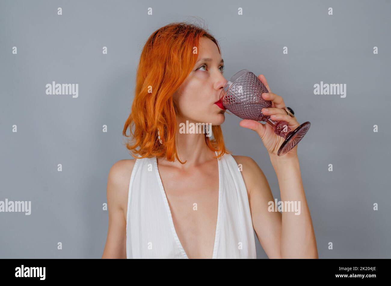 Porträt einer rothaarigen Frau in Weiß mit einem Glas Wein. Eine Frau trinkt aus Glas. Seitenansicht. Stockfoto