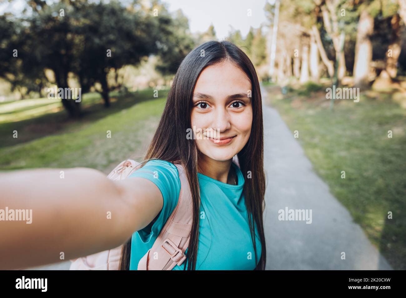 Lächelndes junges Mädchen mit türkisfarbenem T-Shirt und Kopfhörern, das mit ihrem Handy ein POV-Selfie an einem natürlichen Ort macht Stockfoto