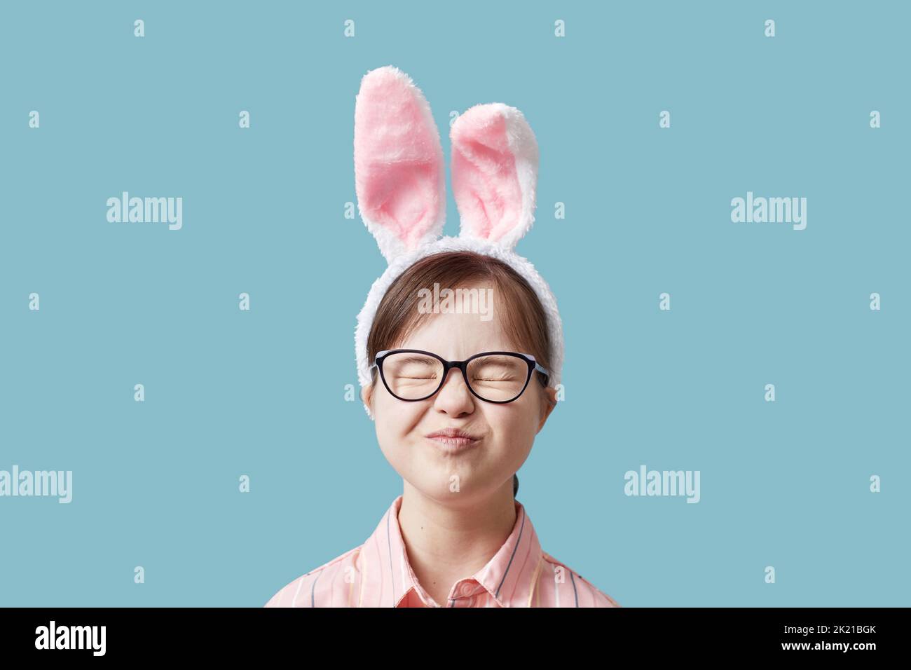 Nettes Mädchen mit Down-Syndrom trägt Osterhasen Ohren vor blauem Hintergrund und macht Gesichter Stockfoto