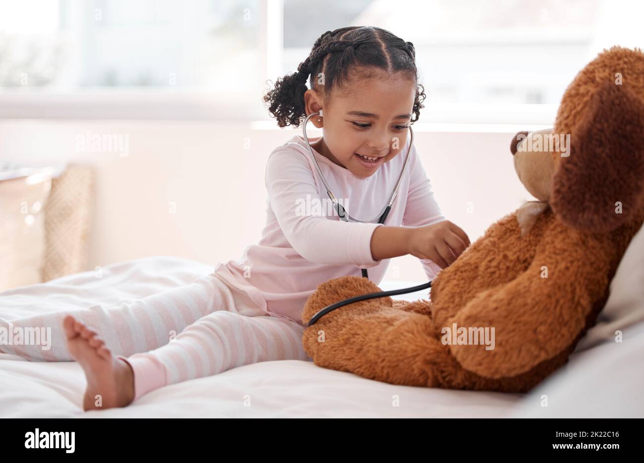 Ein schwarzes Mädchen spielt einen Krankenhausarzt mit einem Teddybären auf ihrem Bett, hält ein Stethoskop und stellt sich ein Kind vor. Entwicklung von Kindern für einen zukünftigen Job als Stockfoto