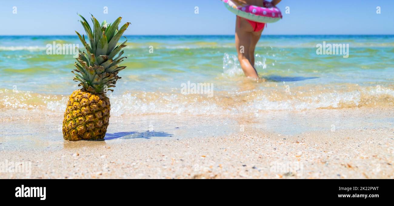 Ananas am Strand und Kind läuft im Wasser Hintergrund. Tropischer Hintergrund. Sommerferienkonzept. Hochwertige Fotos Stockfoto