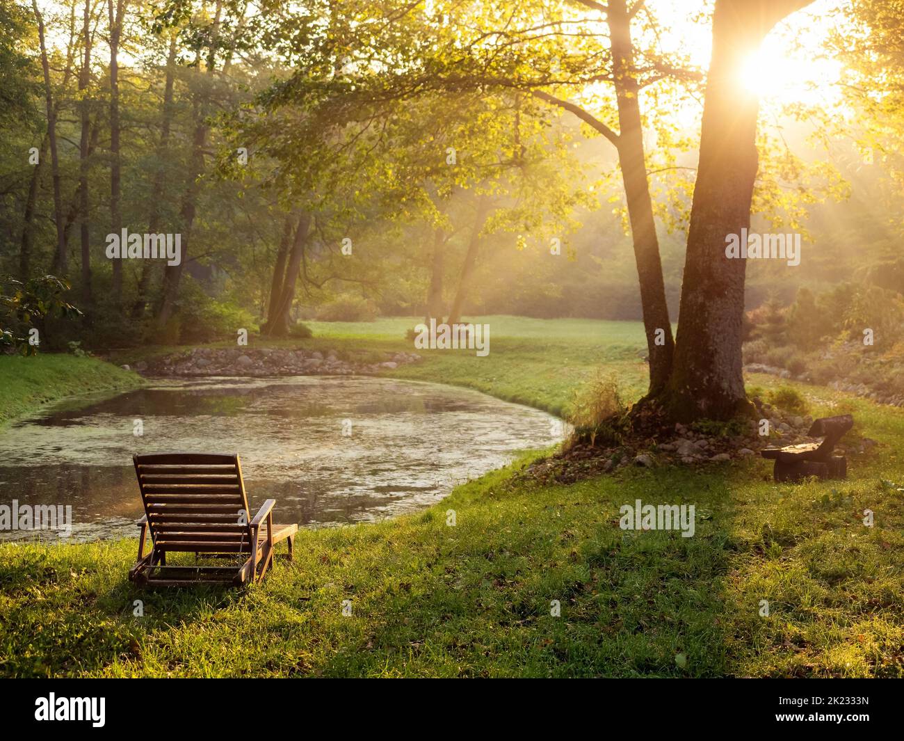 Alter Holzdeckstuhl in einem Herbstgarten mit einem Teich in einem beleuchteten Sonnenuntergang Licht, ruhige herbstliche Stimmung Stockfoto