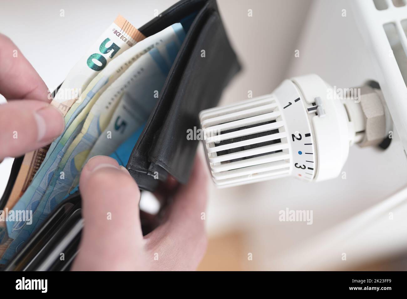 Nahaufnahme der Person, die ihre Brieftasche mit Bargeld neben dem heruntergedrehten Thermostat am Heizkörper hält, Konzept für steigende Energie- und Heizkosten Stockfoto