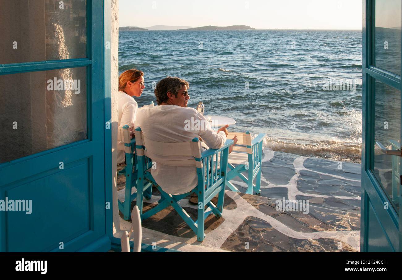 Ein junges Touristenpaar, das in einer Bar am Wasser auf Mykonos, Griechenland, sitzt und den Sonnenuntergang beobachtet Stockfoto