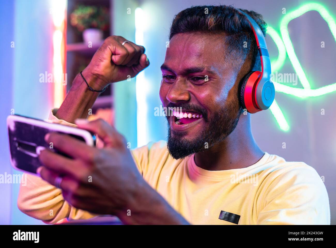 Begeisterter Gamer mit Kopfhörern feiert das Gewinnspiel, indem er die Hände hebt, während er zu Hause ein Live-Videospiel auf dem Mobiltelefon spielt - Konzept des Erfolges Stockfoto