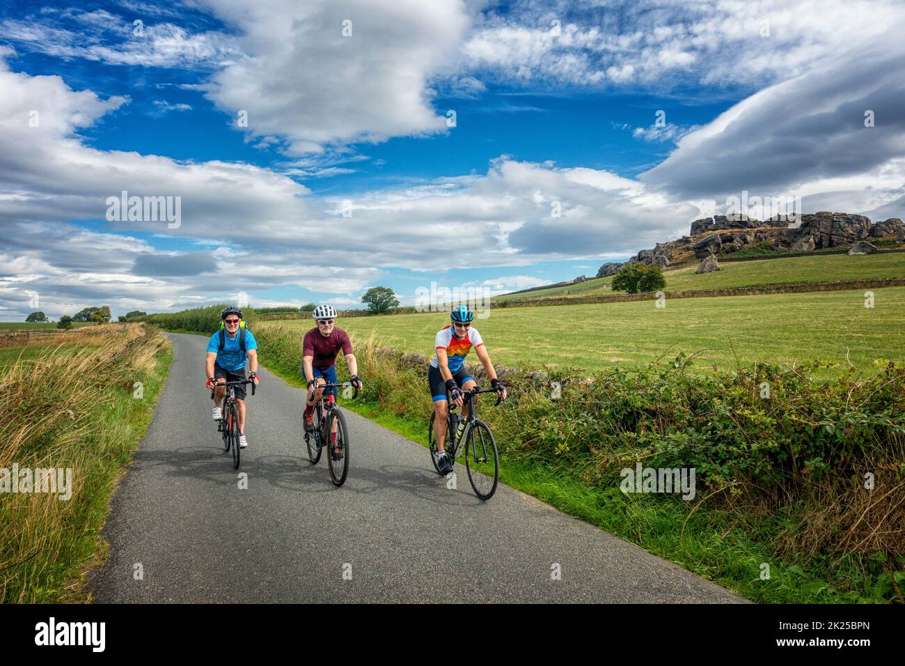Drei männliche Radfahrer treffen sich auf dem Fahrrad auf einer Landstraße mit Almscliffe Crag, North Yorkshire, England, Großbritannien Stockfoto