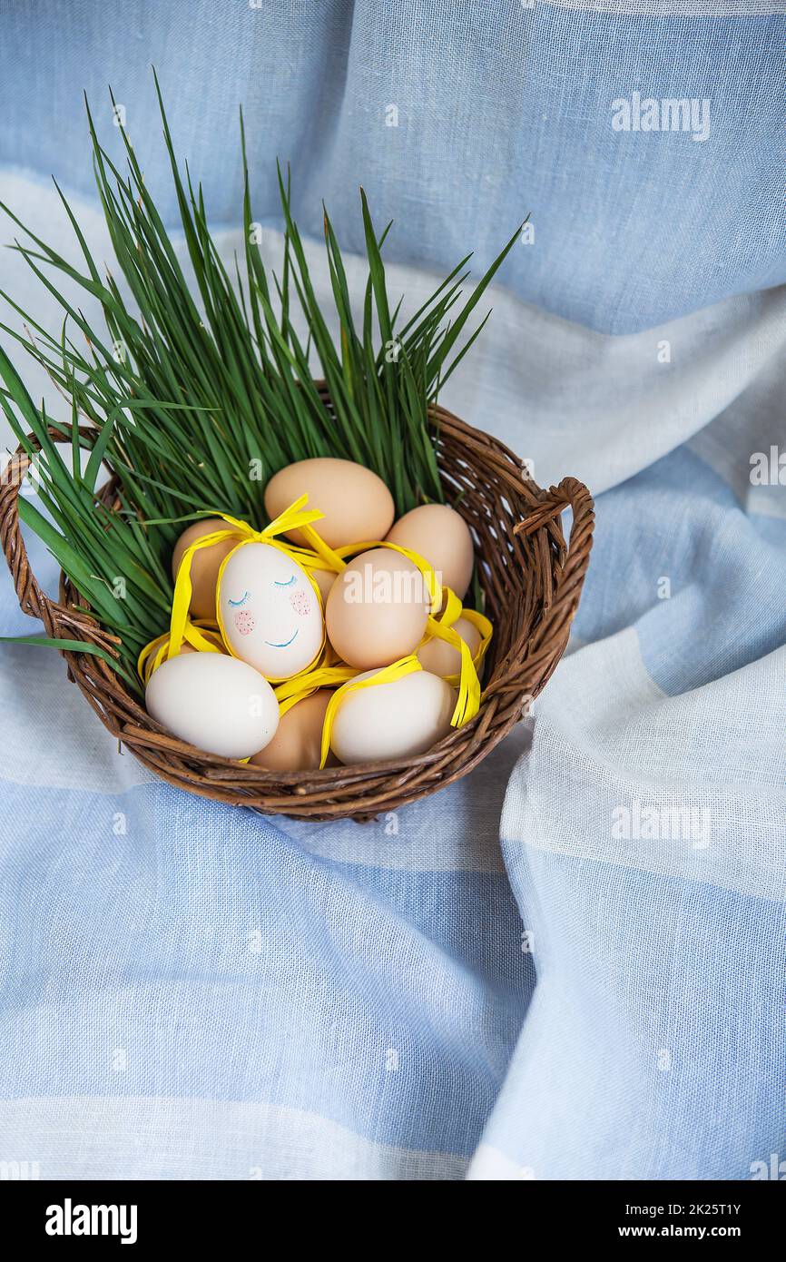 Bemalte Ostereier, ein Ei mit einem süßen Gesicht, Eier liegen in einem Holzkorb zusammen mit grünem Gras. Postkarte für Ostern. Stockfoto