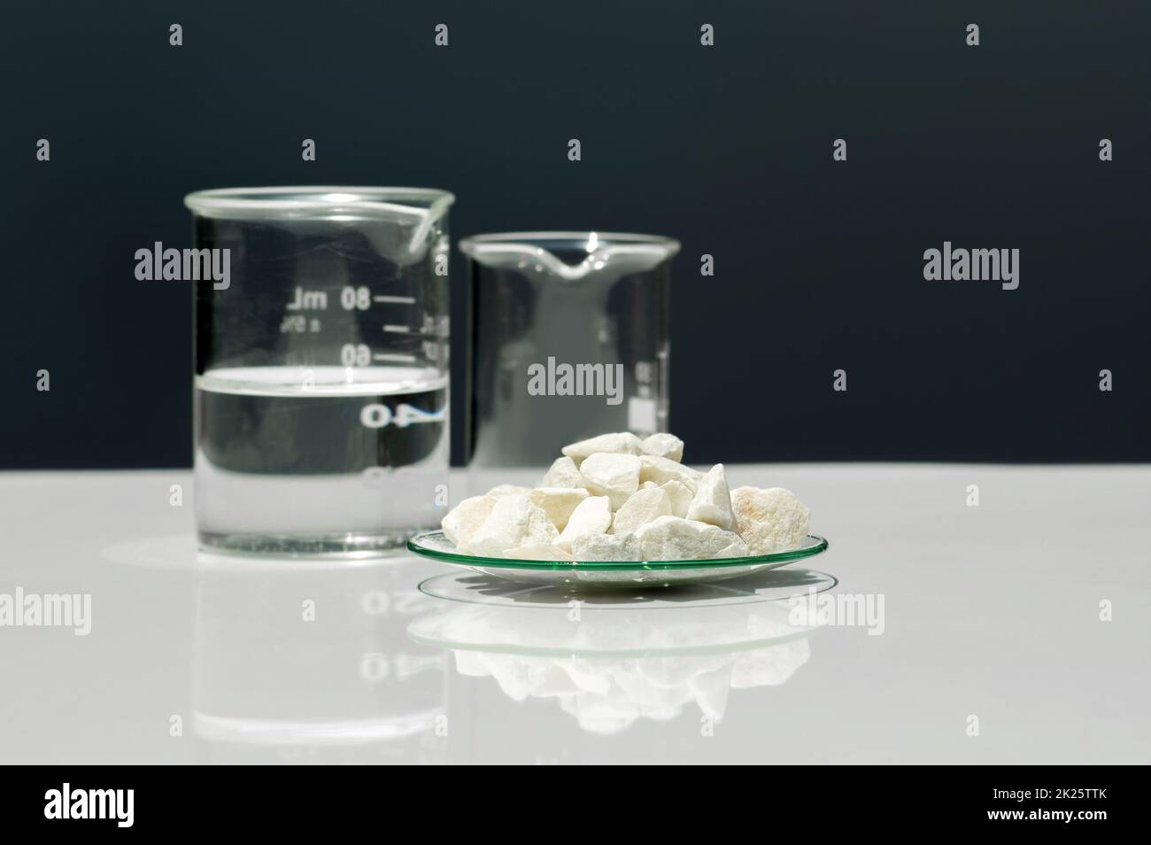 Kalziumkarbonat-Chip in chemischem Uhrglas neben kristallklarer Flüssigkeit im Becher. Chemische Inhaltsstoffe auf weißem Labortisch. Seitenansicht Stockfoto