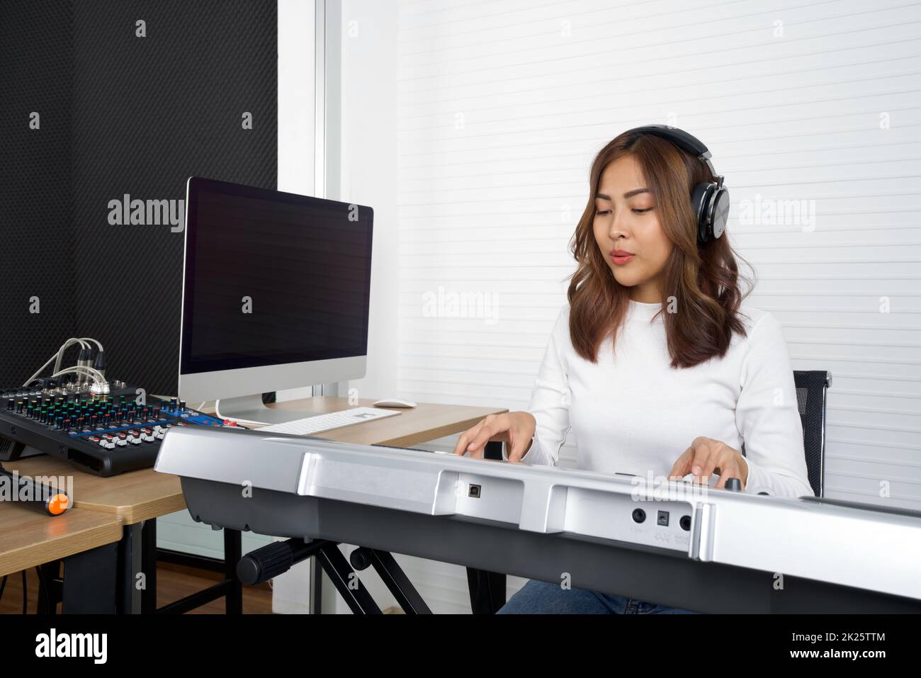 Junge asiatische Frau mit Kopfhörern singen, während sie eine elektrische Tastatur spielt. Musiker, die Musik im professionellen Aufnahmestudio produzieren. Stockfoto