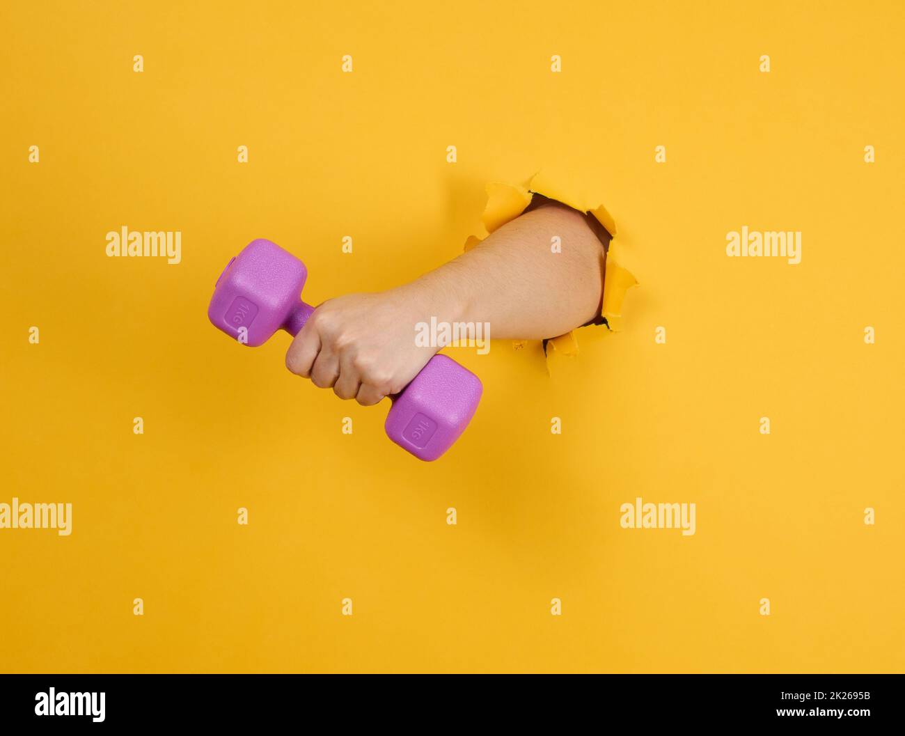 Weibliche Hand hält eine Plastik-Kilogramm-Kurzhantel auf gelbem Hintergrund, ein Teil des Körpers ragt aus einem gerissenen Loch auf einem Papierhintergrund heraus. Gesunder Lebensstil, Sport Stockfoto