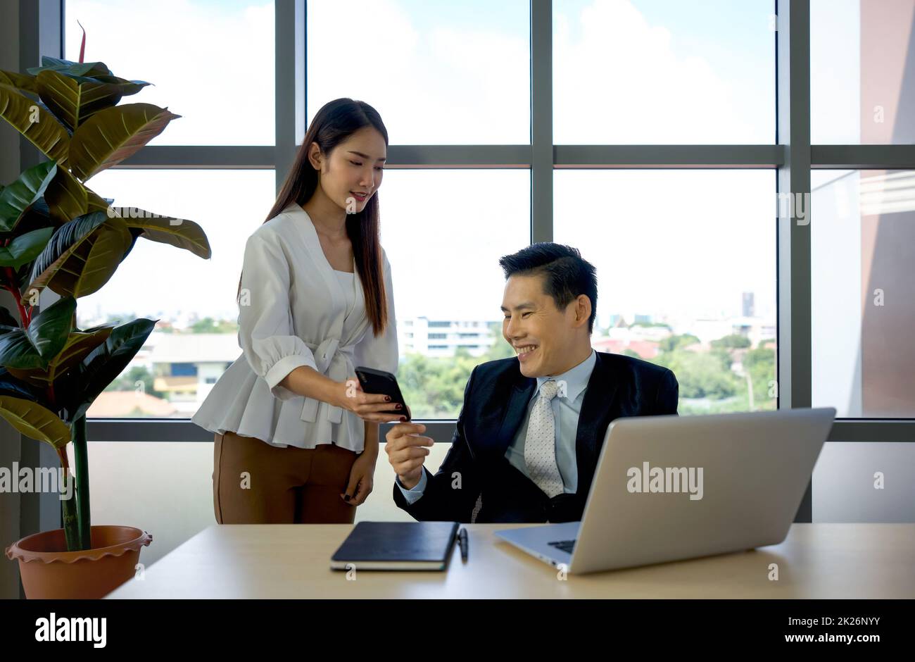 Die junge asiatische Sekretärin zeigt den täglichen Terminplan vom Smartphone bis zum Geschäftsführer in Anzug. Morgendliche Arbeitsatmosphäre in einem modernen Büro. Stockfoto