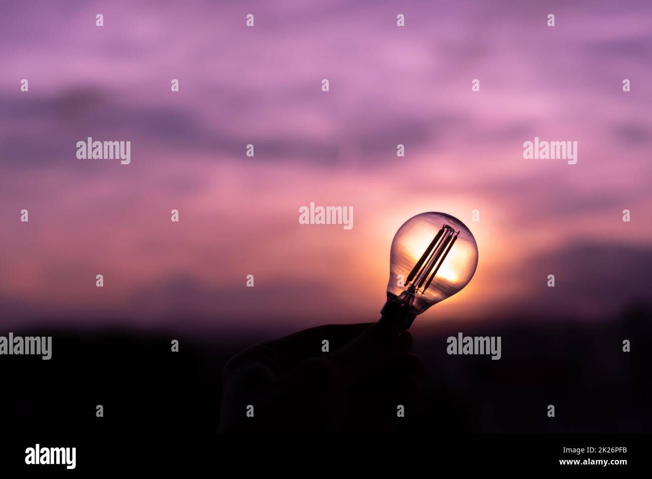 Silhouette einer Glühbirne, die von der Hand einer Person in einem violetten Sonnenuntergangshimmel gehalten wird, der sprachlos bleibt Stockfoto