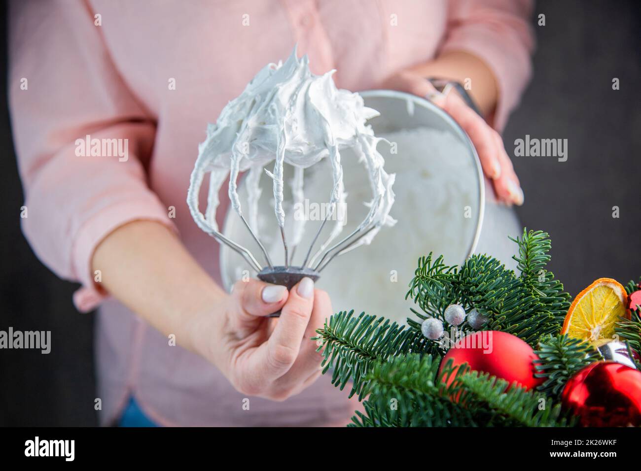 Der Schneebesen eines elektrischen Mischers in einer weißen Masse aus Baiser mit stehenden Gipfeln wird in der Hand eines Konditors gehalten, vor dem Hintergrund einer Eisenkammer, die Zweige eines Weihnachtsbaums und Silvesterspielzeug sind von der Seite sichtbar. Stockfoto