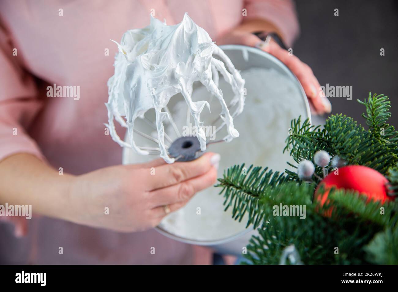Der Schneebesen eines elektrischen Mischers in einer weißen Masse aus Baiser mit stehenden Gipfeln wird in der Hand eines Konditors gehalten, vor dem Hintergrund einer Eisenkammer, die Zweige eines Weihnachtsbaums und Silvesterspielzeug sind von der Seite sichtbar. Stockfoto