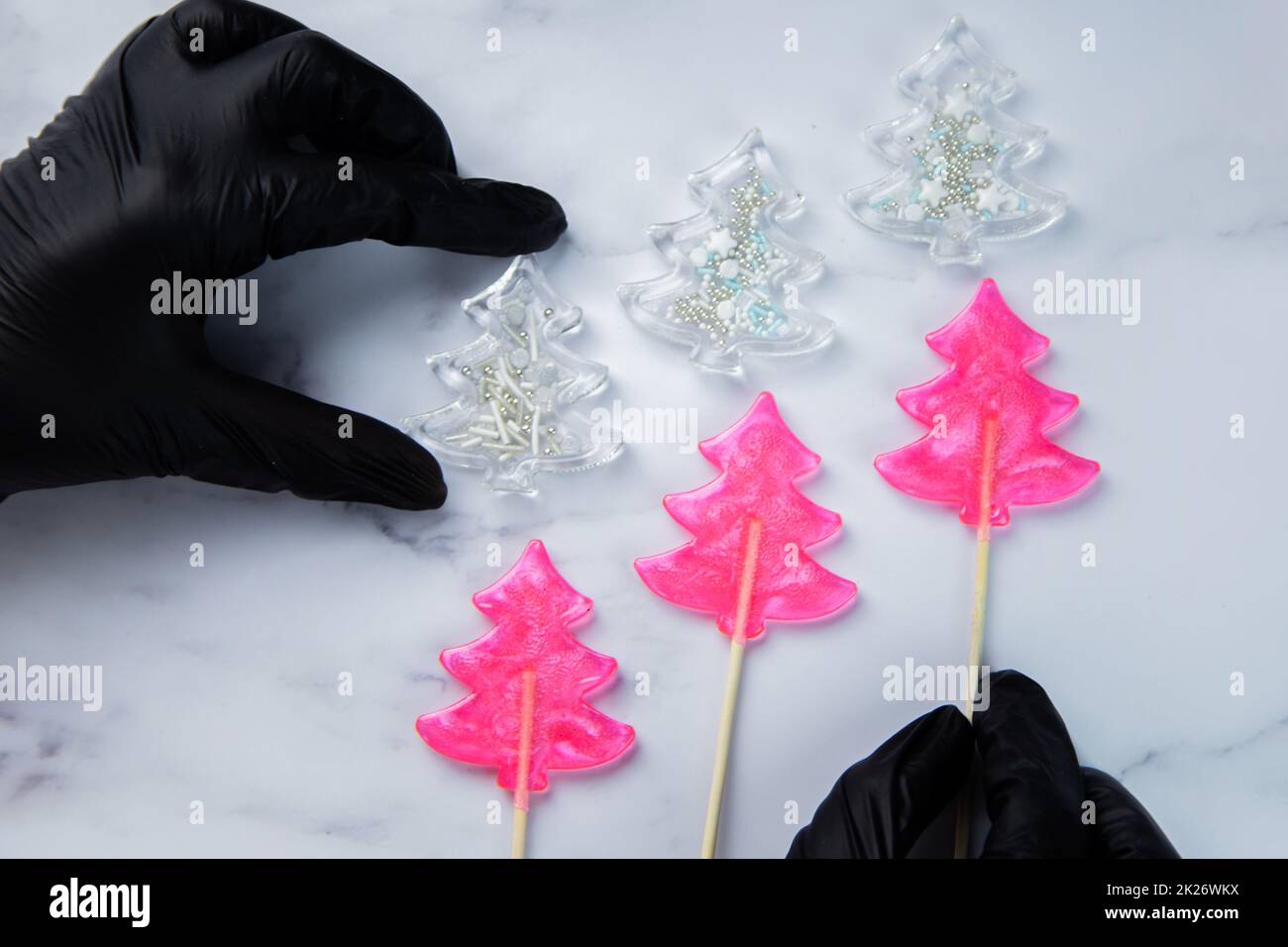 Drei pinkfarbene Lutscher in Form von Weihnachtsbäumen liegen auf weißem Marmor, über ihnen sind weiße Weihnachtsbäume - Lutscher mit Pulver, die extremen halten Hände in schwarzen Handschuhen. Stockfoto