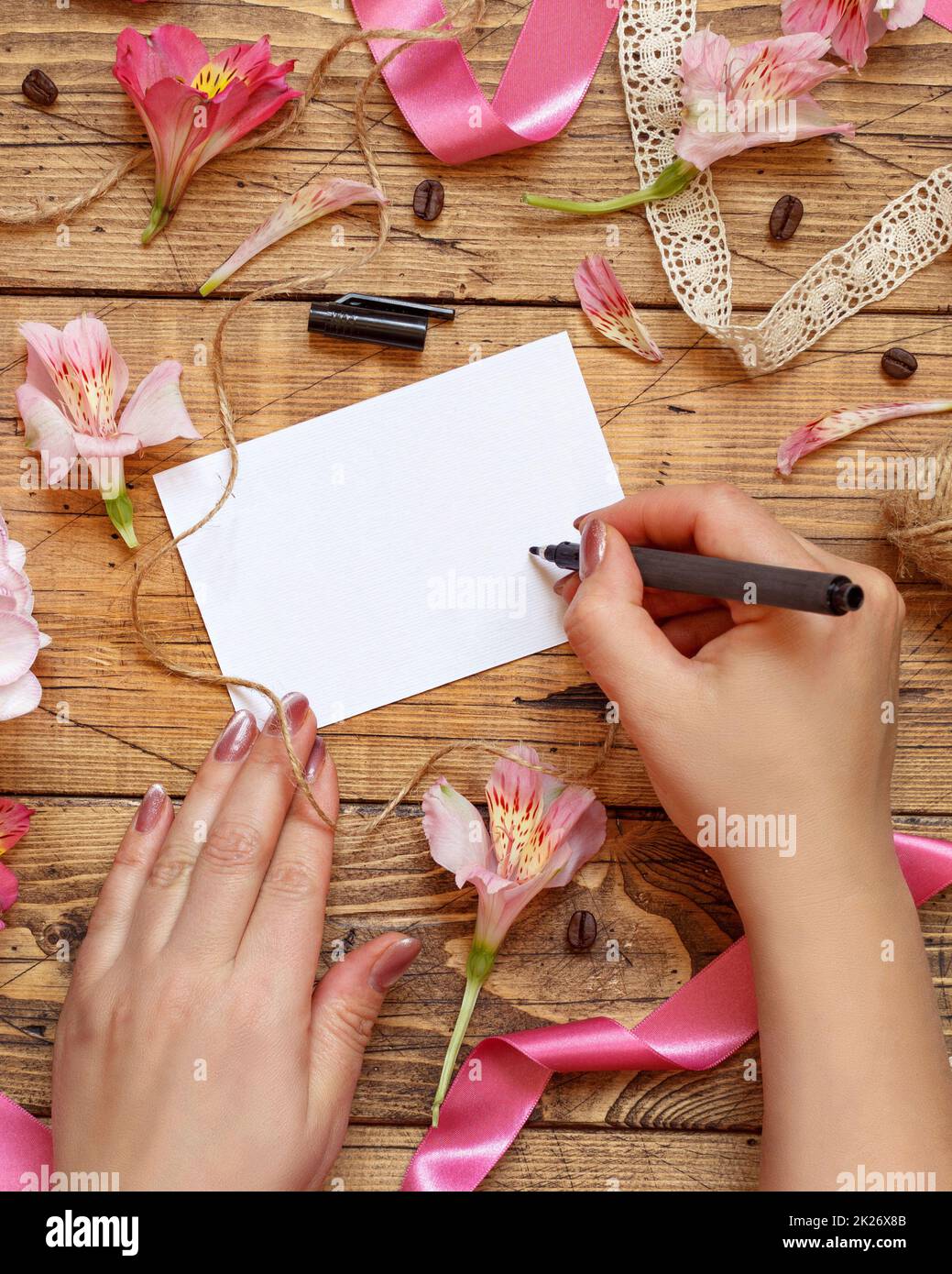 Die Hände schreiben eine Karte auf einen Holztisch zwischen rosa Blumen Stockfoto
