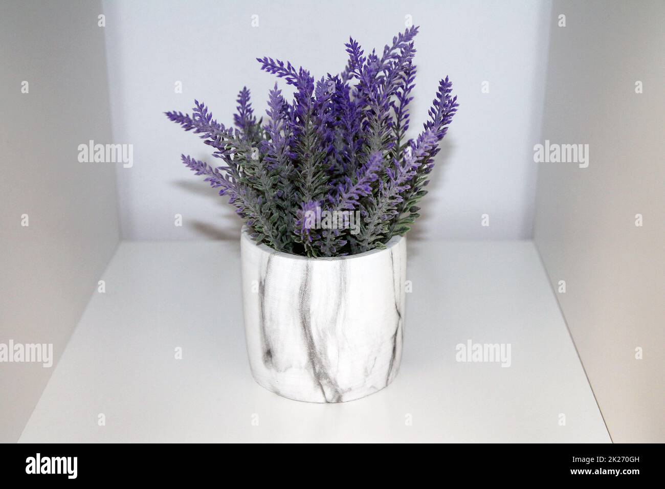 Eine violette und grüne künstliche Lavendelpflanze mit Blumen und eine Marmorvase auf einem weißen Regal Stockfoto