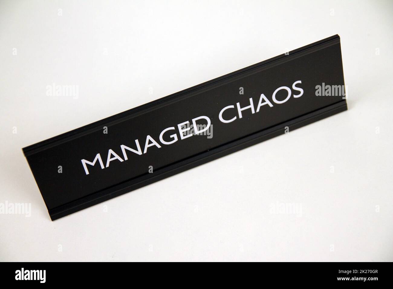 Verwaltetes Chaos, schwarzes und silbernes Metallschild auf einem weißen Regal Stockfoto